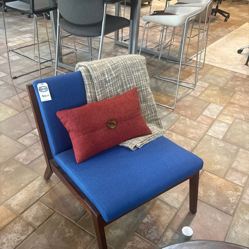 Blue upholstered chair with wooden frame in a showroom, accented by a red pillow and gray throw; tag hanging from the back. Background shows tables and white chairs.