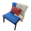 Blue upholstered lounge chair with wooden legs, accented by a red lumbar pillow and a gray woven throw draped over the back as a cozy seating setup.