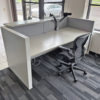Empty office cubicle with an L-shaped light-gray desk, gray divider panels, and a swiveling office chair by a large window.