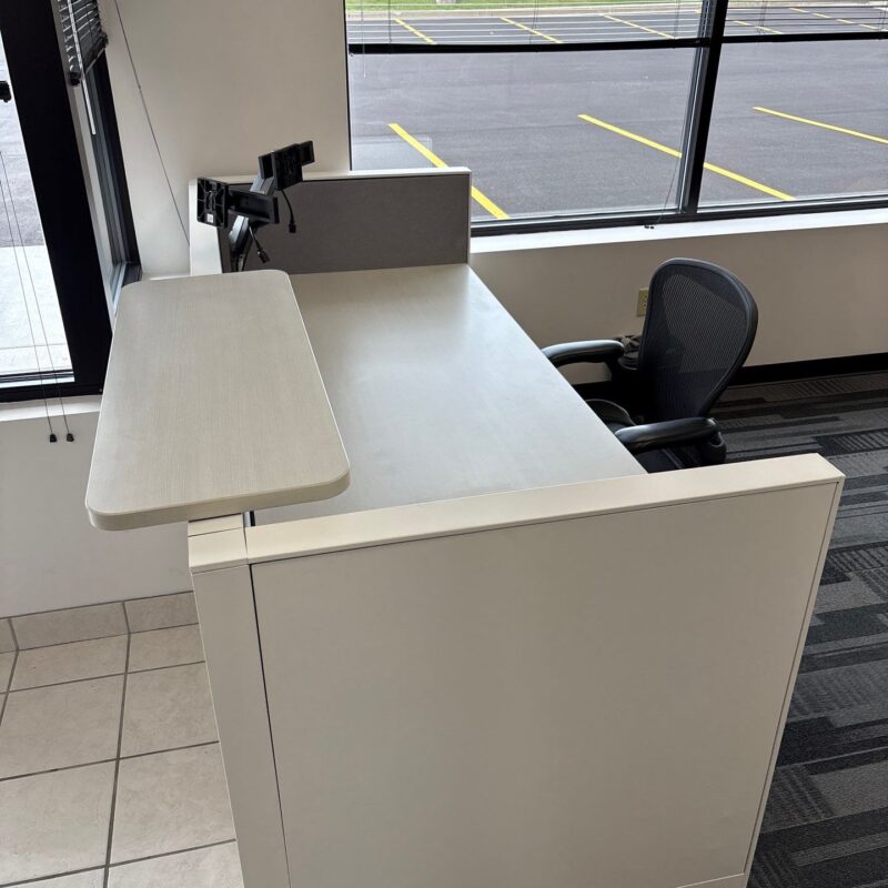 Workstation with a white reception desk and pull-out counter, black mesh chair, monitor arms, near large windows overlooking a parking lot and green lawn.