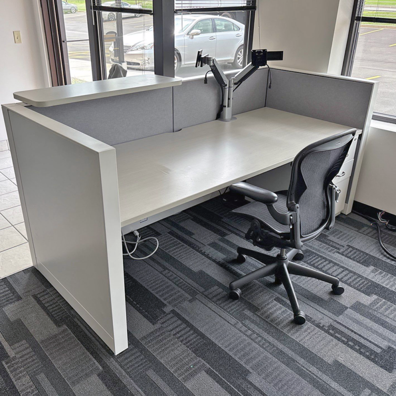 Empty office cubicle with an L-shaped light-gray desk, gray divider panels, and a swiveling office chair by a large window.
