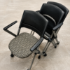 Stacked black office chairs with perforated backrests, metal frames, and rolling bases on a tiled floor.