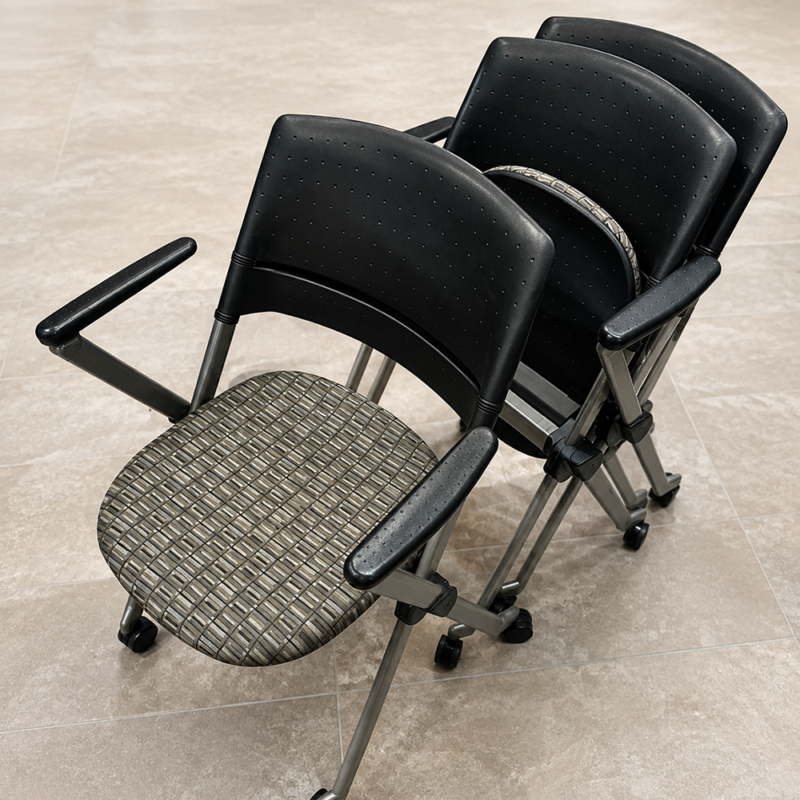 Stacked black office chairs with perforated backrests, metal frames, and rolling bases on a tiled floor.