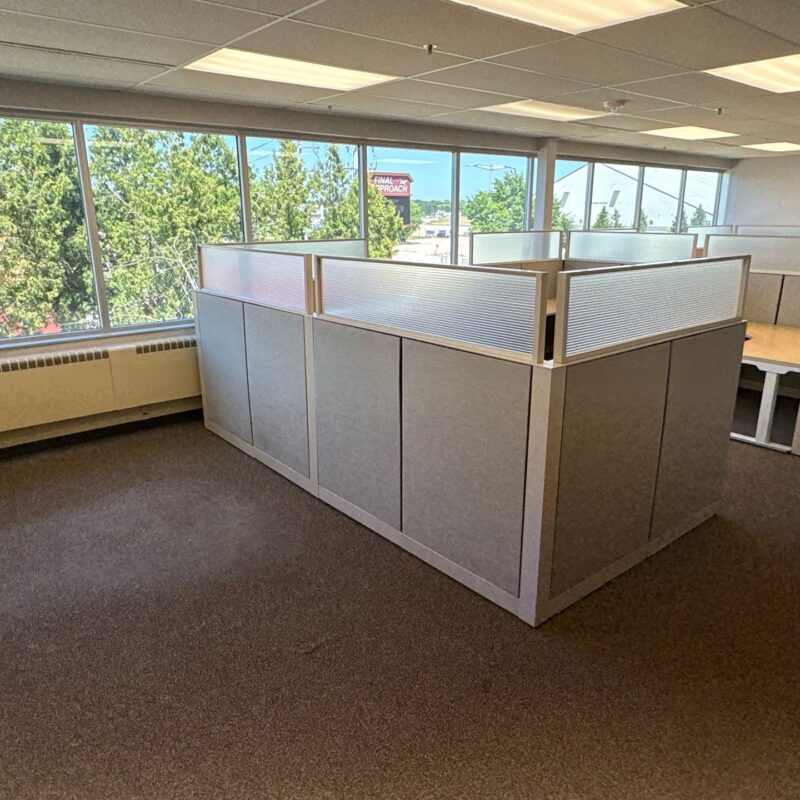 Open-plan office with gray cubicles along large windowed wall, trees outside, and a wall-mounted TV in the background.