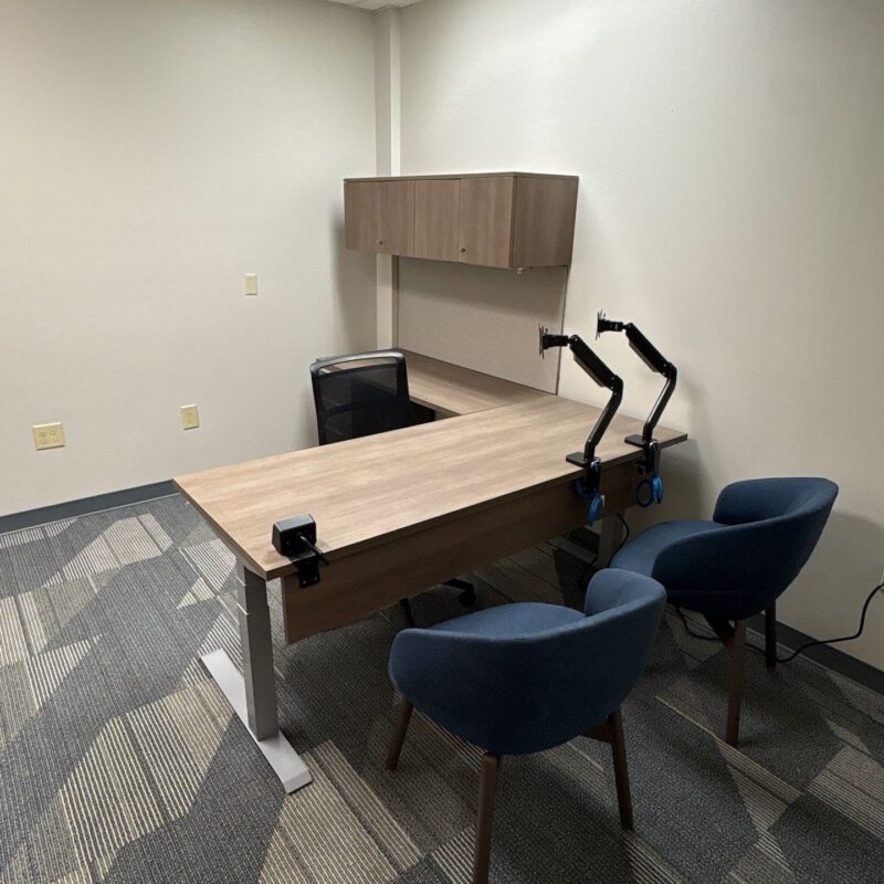 Office study room with a long wooden desk, black mesh chair, wall cabinet, and two adjustable desk lamps; blue visitor chairs around the desk.