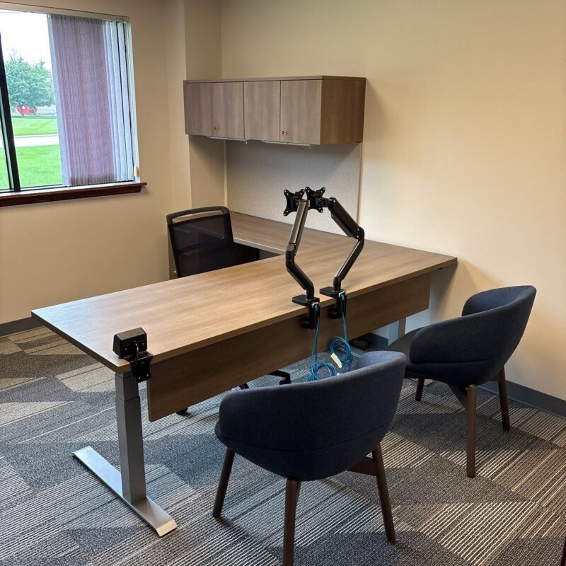 Modern office workspace with a long desk, two adjustable monitor arms, and two blue chairs facing a wall with cabinets above.