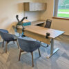 Office workspace with a long wooden desk, two blue chairs, wall cabinets, and adjustable monitor arms on the desk.