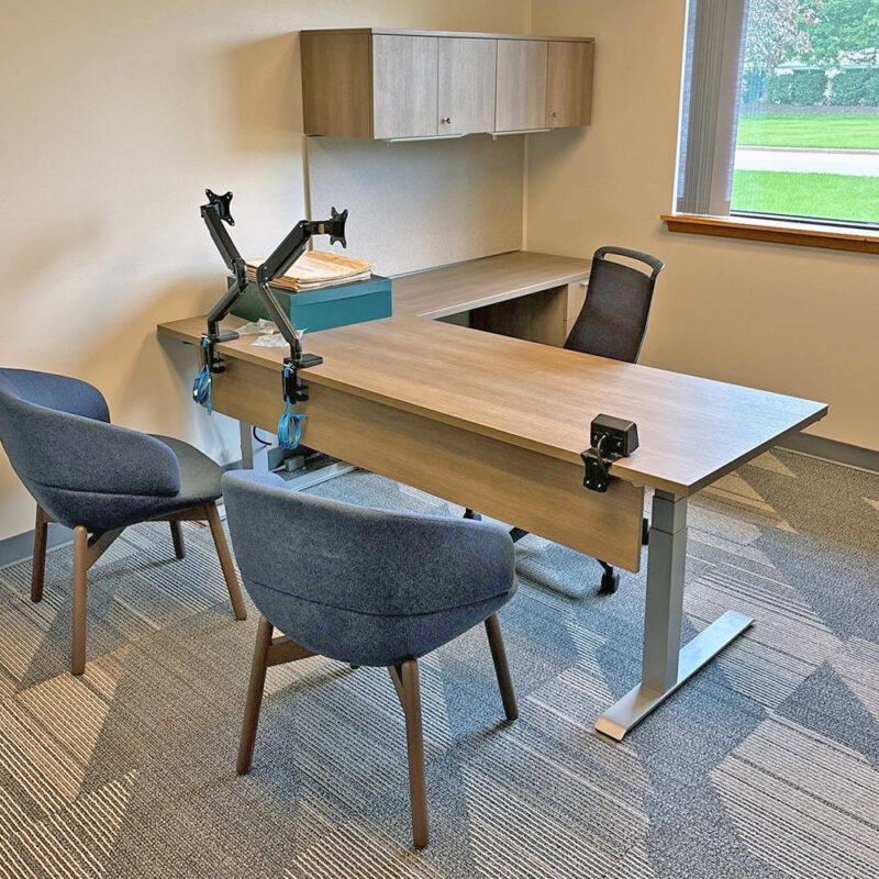 Office workspace with a long wooden desk, two blue chairs, wall cabinets, and adjustable monitor arms on the desk.