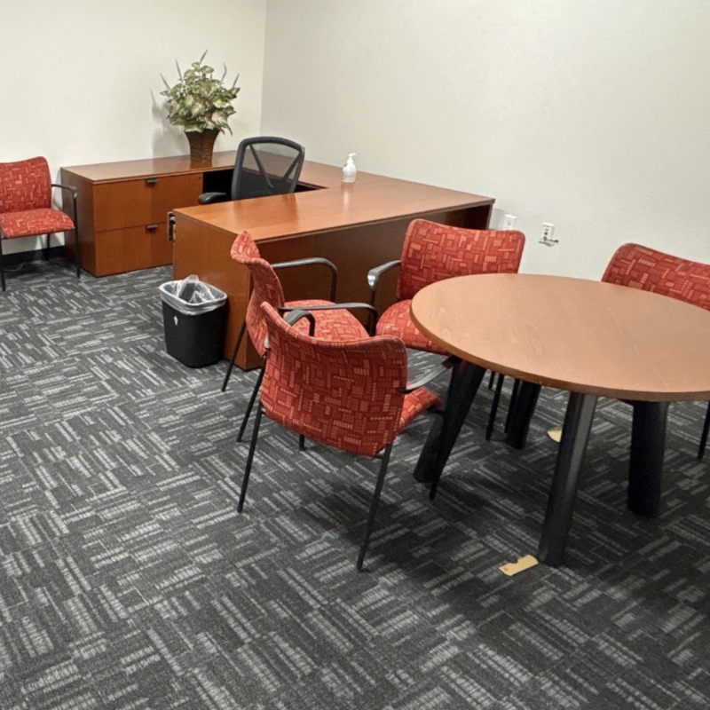Office reception area with wooden desk, black chair, red patterned chairs, and a round table nearby