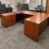 L-shaped wooden office desk with a small drawer cabinet on the left and a hole for cable management on the top surface in a softly blurred office background.