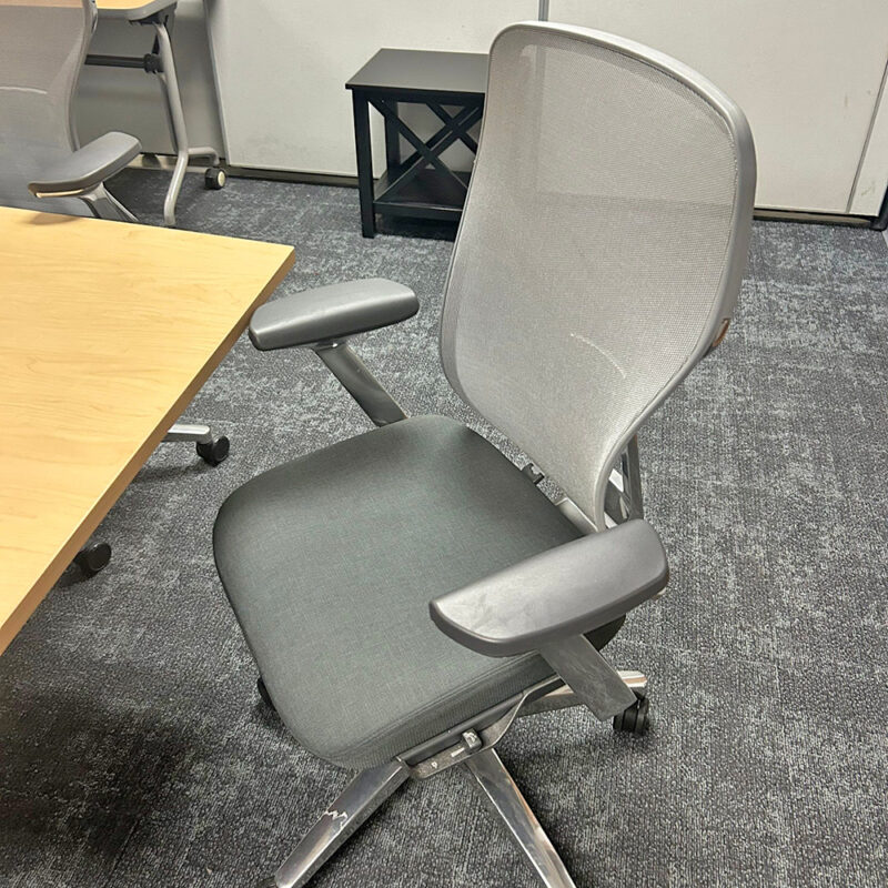 Ergonomic gray office chair with mesh back and padded seat on a wheeled base beside a wooden table in a carpeted room.