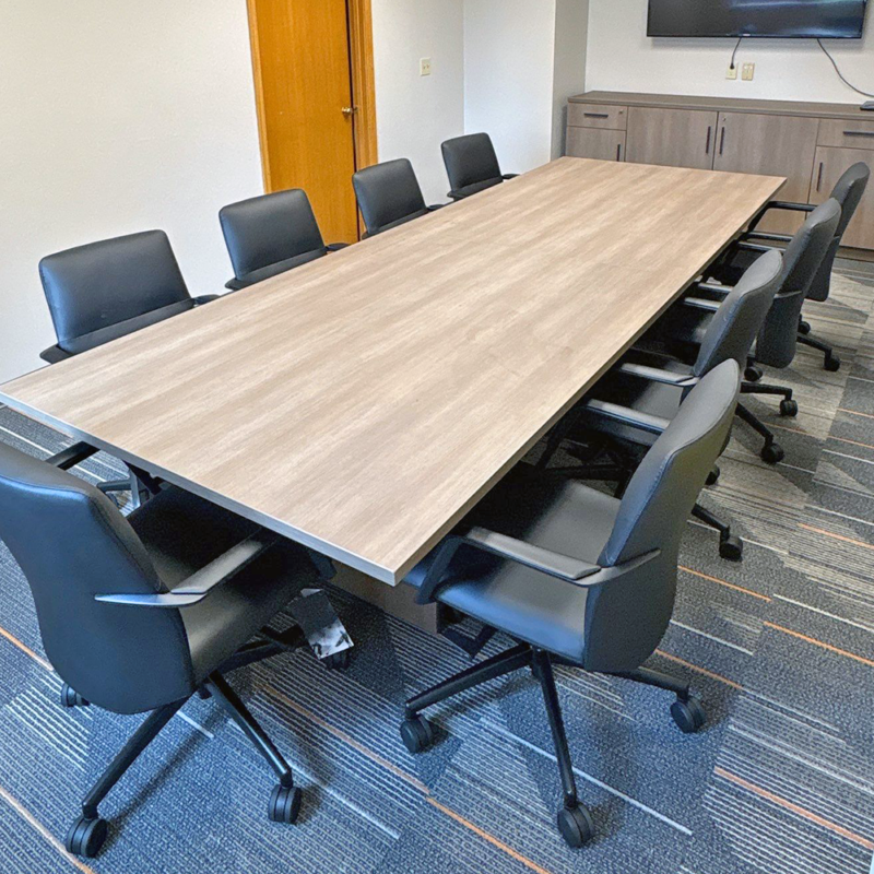 Long wooden conference table surrounded by black rolling chairs in a meeting room, with a cabinet and TV on the back wall.