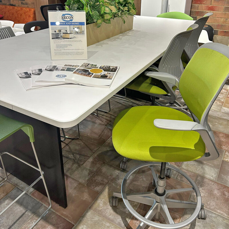 Modern office conference area with a lime-green swivel chair at a long white table, brochures scattered nearby and a green plant centerpiece.
