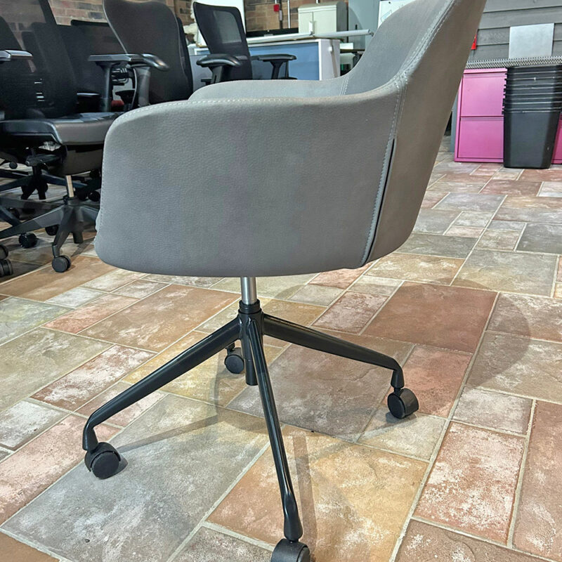 Gray upholstered office chair with a five-star wheeled base on a tiled showroom floor, with other chairs and office furniture in the background.