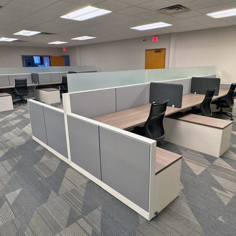 Open-plan office with grey cubicle partitions, wooden desks, and black ergonomic chairs in a spacious workspace.