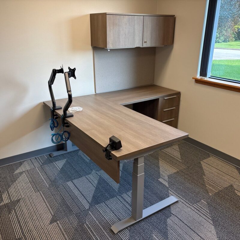 Modern L-shaped office desk with a monitor arm and wall cabinet, near a window with a grassy outdoor view.