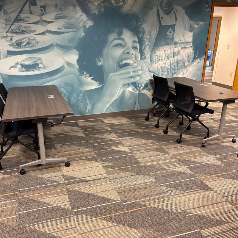 Two rectangular tables with rolling chairs in a meeting area, backed by a large blue-toned mural of a woman taking a big bite of a burger.