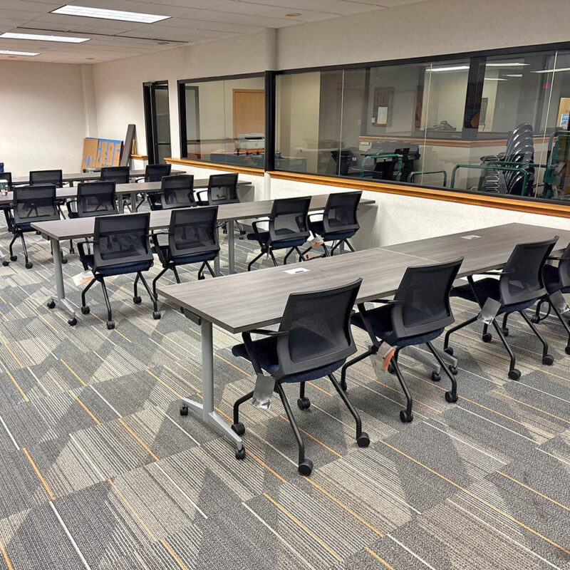 Empty training room with gray rectangular tables and black rolling chairs arranged in rows, projector screen at front