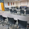 Modern classroom or training room with multiple gray tables and black rolling chairs arranged in rows, a projector screen at the front, and posters on the side wall