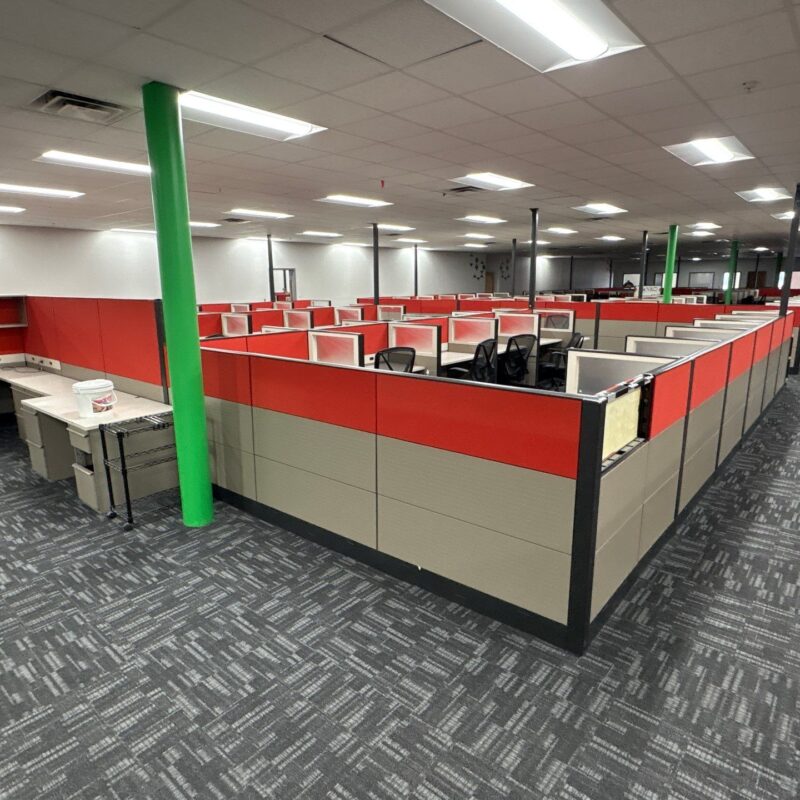 Open-plan office with red cubicle partitions, gray desks, and black chairs, supported by green columns under a grid ceiling with fluorescent lights, creating an empty workspace