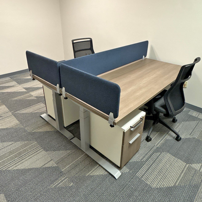 Two-person office workstation with blue fabric privacy partitions, wooden desks, and a mesh-backed chair in a neutral room.