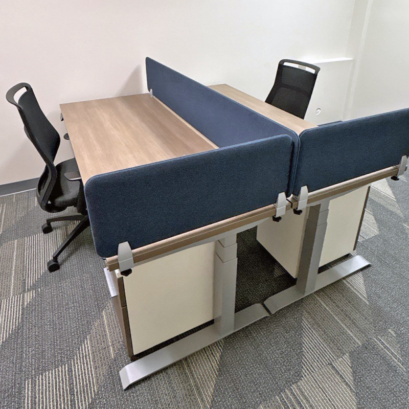 Shared two-person desk with blue fabric privacy panels and two black rolling office chairs in a modern office setting.