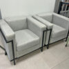 Two light gray upholstered armchairs with metal frame legs joined by a small divider, arranged along a wall in a showroom.