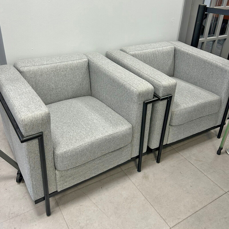 Two light gray upholstered armchairs with metal frame legs joined by a small divider, arranged along a wall in a showroom.