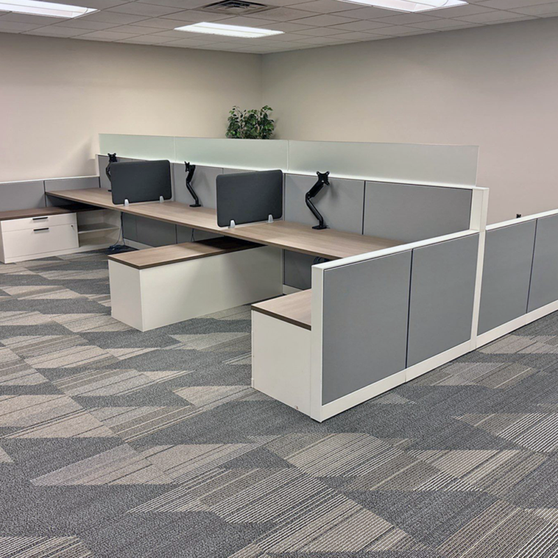 Open-plan office with row of desks and grey divider panels, adjustable monitor arms, and a small plant in the back corner.