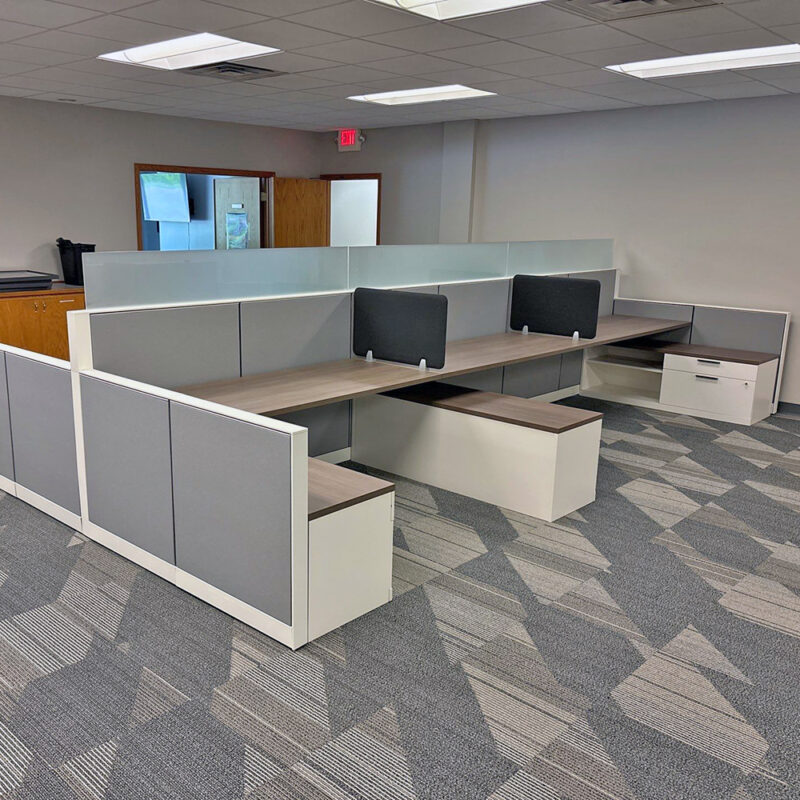 Empty office cubicle area with L-shaped desks, gray privacy panels, and frosted glass dividers in a neutral-lit workspace