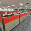 Open-plan office with rows of red-and-gray cubicles and empty workstations under bright fluorescent lights.