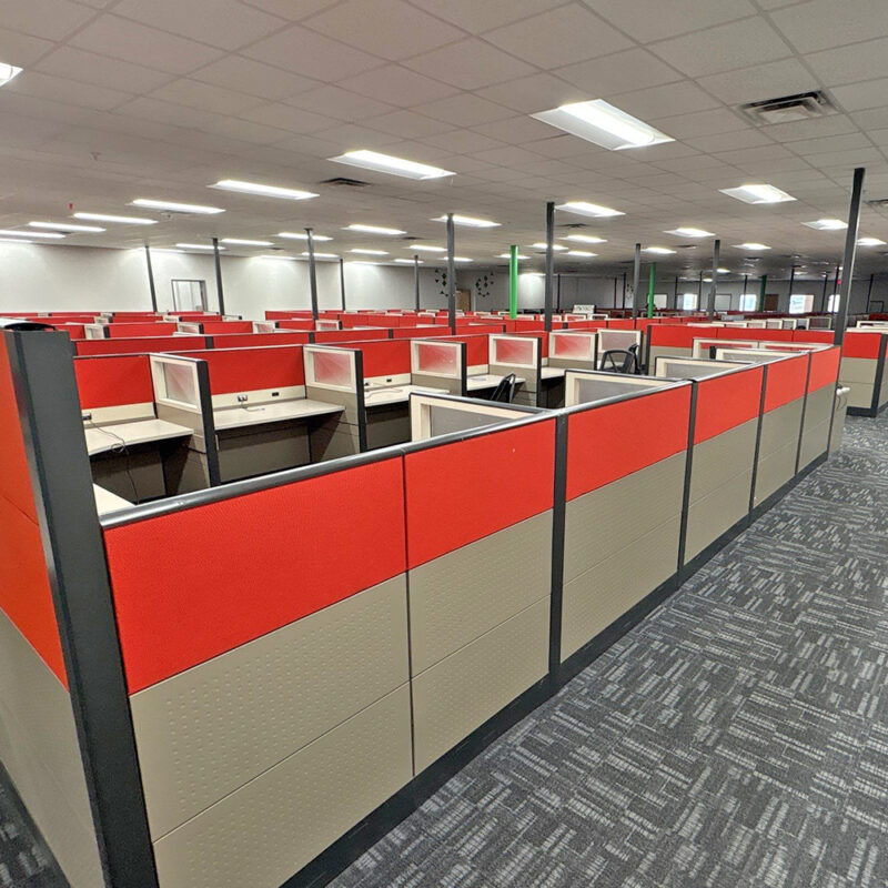 Open-plan office with rows of red-and-gray cubicles and empty workstations under bright fluorescent lights.